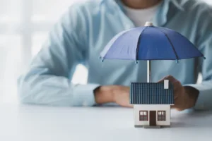 Tiny toy house on a desk with an umbrella. Man in a blue button down is sitting behind the house out of focus.